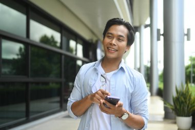 Neşeli genç Asyalı adam modern bir binada yürüyor ve akıllı telefonunu kullanıyor..