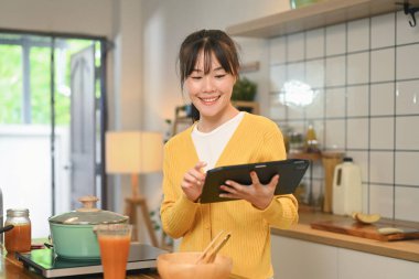 Mutfak tezgahında durup yemek tarifine ya da dijital tabletteki yemek tariflerine bakan gülümseyen genç bir kadın..