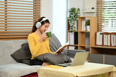 Kulaklıklı genç Asyalı kadın sıcak çay içiyor ve evdeki kanepede kitap okuyor..