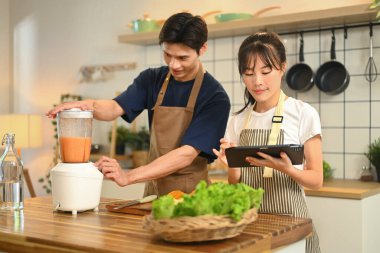 Mutlu genç çift sağlıklı meyve suları hazırlıyor ve dijital tablette online bir tarifi takip ediyor..