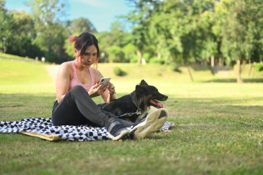 Antrenmandan sonra parktaki bir battaniyede köpeğiyle otururken cep telefonu kullanan genç bir kadın..