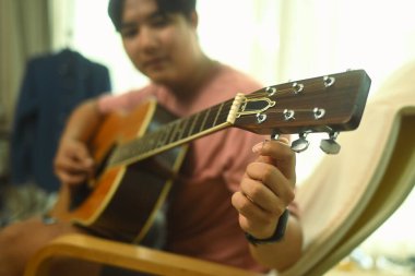 Rahat bir oturma odasında akustik gitar çalan sıradan giysili Asyalı genç bir erkek..