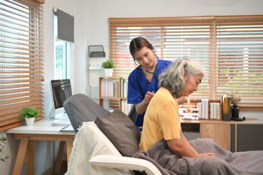 Mavi önlüklü bir sağlık uzmanı, yaşlı bir kadın hastanın nefes alışını kontrol etmek için steteskop kullanıyor..