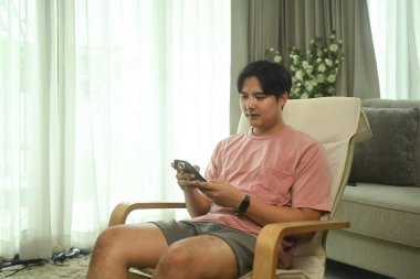 Evdeki koltukta cep telefonu kullanan gülümseyen Asyalı genç adam..