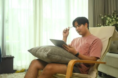 Asyalı erkek, evdeki minderli sandalyede dijital tabletle çalışıyor..