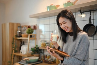 Büyüleyici genç Asyalı kadın mutfakta süt içip kitap okuyor..