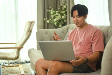 Asyalı serbest çalışan, günlük giysiler içinde, video görüşmesi için laptop kullanıyor, evdeki kanepede oturuyor..