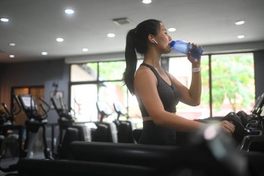 Spor salonunda koşu bandında çalışırken su içen genç bir fitness kadını..