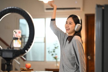 Kulaklıklı neşeli genç bir kadın akıllı telefonuyla içeriğini kaydediyor. Tripoda takılı halkalı bir lambayla..