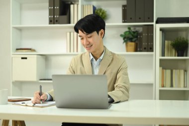 Dizüstü bilgisayar kullanan ve büro masasında notlar yazan modaya uygun takım elbiseli genç işadamı..