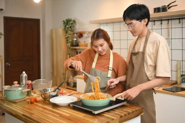 Güzel genç bir kadın yeşil bir tavada spagetti pişirirken kocası da onun yanında yemek pişirmesine yardım ediyor..