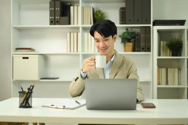 Başarılı genç işadamı kahve içiyor ve bilgisayarla ofiste çalışıyor..