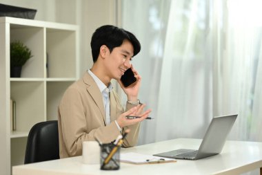 Gülümseyen erkek finans danışmanı cep telefonuyla konuşuyor ve ofiste dizüstü bilgisayar kullanıyor..