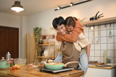 Sıcak bir mutfakta yemek pişiren önlük giyen neşeli genç çift. İlişkiler, yemek pişirme ve mutlu bir ev hayatı..