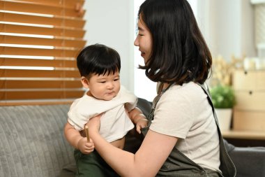 Gülümseyen genç bir anne kucağında bebeğiyle evdeki kanepede oturuyor. Çocukluk ve bakım konsepti.