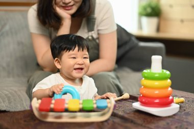 Sevimli erkek bebek yerde oturuyor, renkli oyuncaklarla oynuyor ve anne çocuğunu sevgi dolu bir ifadeyle seyrediyor..