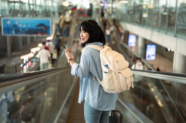 Havaalanı terminalinde yürüyen merdivende cep telefonu kullanan mutlu, genç bir kadın..