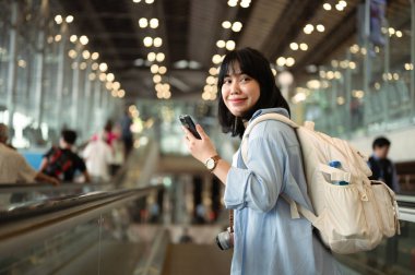 Havaalanı terminalinde yürüyen merdivende cep telefonu kullanan mutlu, genç bir kadın..