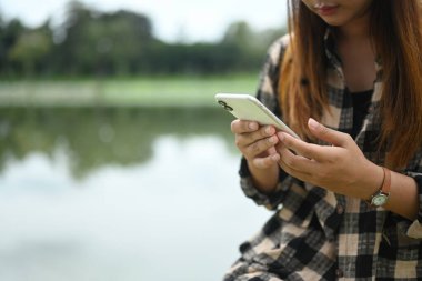 Sakin bir nehrin kenarında dinlenirken cep telefonu kullanan kareli tişörtlü bir kadın..
