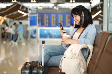 Havaalanı terminalinde bavuluyla oturan ve cep telefonu kullanan gülümseyen genç bayan turist..