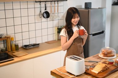 Mutfakta kahvaltı hazırlarken önlük giymiş genç ve çekici bir kadın sıcak bir içkinin tadını çıkarıyor..