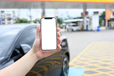 Benzin istasyonunda bir arabanın önünde boş beyaz ekranlı akıllı telefonu tutan eller..