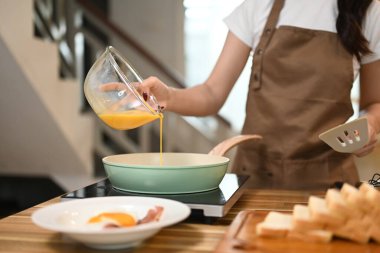 Önlüklü genç bir kadın cam kaseden çırpılmış yumurtaları kızartma tavasına döküyor..