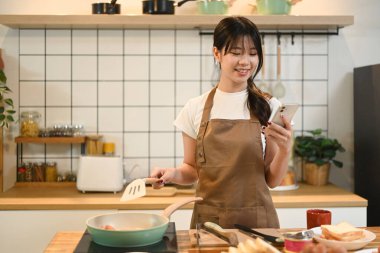 Ahşap bir atölyede yemek hazırlarken akıllı telefon kullanan gülümseyen genç kadın..