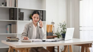 Güzel genç girişimci cep telefonuyla konuşuyor ve masadaki belgeleri inceliyor..