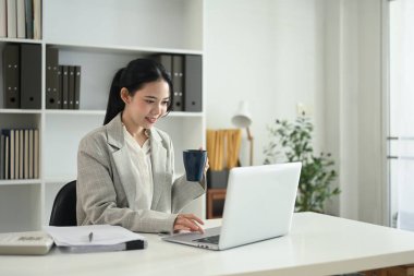 Güzel bir iş kadını elinde kahve fincanıyla ofisinde dizüstü bilgisayar kullanıyor..