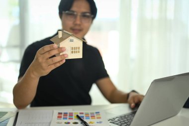 Renk örnekleri ve kumaş örnekleriyle masanda modelin detaylarını incelerken elinde küçük bir model ev tutan adam..