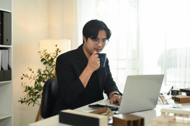 Erkek mimar, masanda model binalar ve malzeme örnekleriyle çalışırken kahvesini yudumluyor..