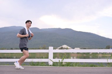 Genç adam kaldırımlı yolda koşuyor manzaralı dağlar ve arka planda sakin bir göl var..