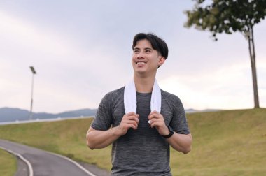 Gülümseyen Asyalı genç adam bulutlu bir günde parkta koşuyor. Fitness ve aktif yaşam tarzı.