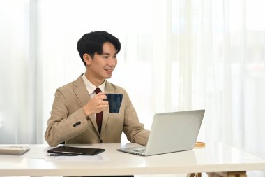 Genç işadamı kahve içiyor ve bilgisayarla parlak ofiste çalışıyor..