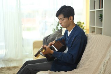 Yakından çekilen adam kanepede gitar çalıyor. Mavi gömlek ve gri pantolon giyiyor.