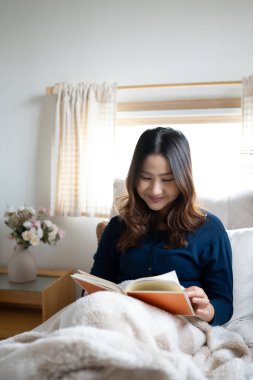 Pencere kenarında kitap okuyan gülümseyen genç bir kadın, yumuşak bir battaniyeye sarılmış..