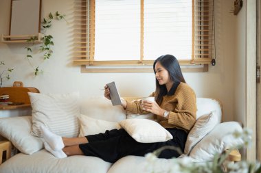 Gülümseyen, kahve içen ve dijital tablet kullanan genç bir kadın..