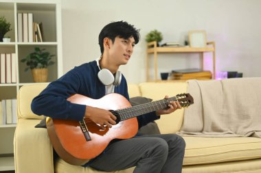 Genç Asyalı adam rahat bir oturma odasında akustik gitar çalışıyor..