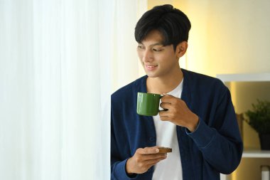 Yakışıklı Asyalı adam pencerede bir fincan kahvenin keyfini çıkarıyor..