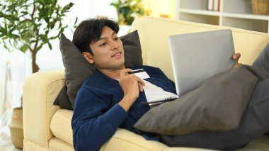 Genç adam online alışveriş yapıyor, kredi kartı tutuyor ve evde dizüstü bilgisayar kullanıyor..