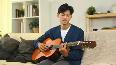 Genç Asyalı adam rahat bir oturma odasında akustik gitar çalışıyor..