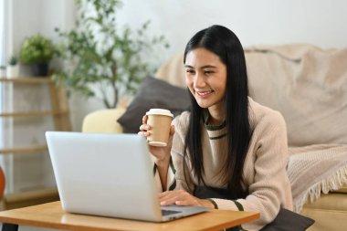 Güzel genç bir kadın, kağıt bardaktan kahve içiyor ve oturma odasında dizüstü bilgisayar kullanıyor..