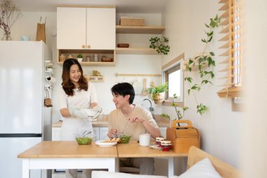 Asyalı genç bir kadın, minimalist bir mutfakta kocasına sağlıklı yemek servisi yapıyor..