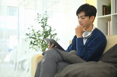 Kanepede otururken dijital tablet kullanan düşünceli genç bir serbest çalışan..