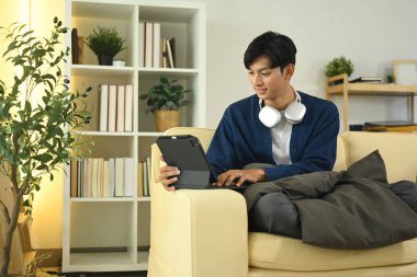 Yakışıklı Asyalı adam dijital tabletle kanepede dinleniyor..