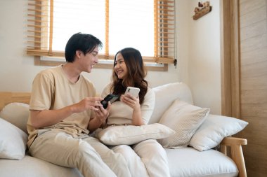 Mutlu Asyalı çift evdeki kanepede akıllı telefon kullanıyor..
