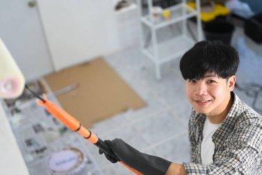 Gülümseyen genç adam duvarları silindirle boyuyor, uzayı dönüştürme sürecinin tadını çıkarıyor..