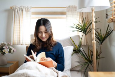 Sakin, genç bir kadın, yumuşak bir battaniyenin altında oturmuş, evdeki pencerenin kenarında kitap okuyor..