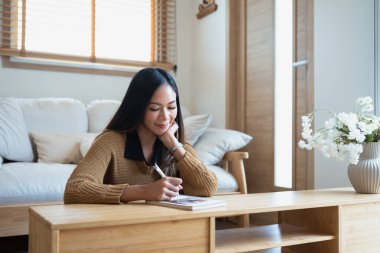 Minimalist bir oturma odasında dinlenirken günlüğe notlar yazan düşünceli bir genç kadın..